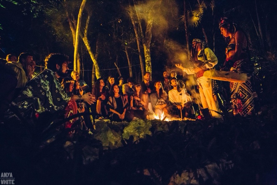 The community gathered around a fire at night in the jungle, listening to two people leading a ceremony.